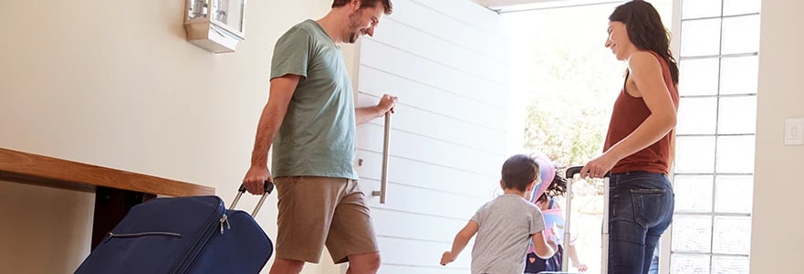 A family of four is preparing to leave their home, with the father pulling a large blue suitcase and the mother guiding their two young children towards the front door, bathed in natural light.